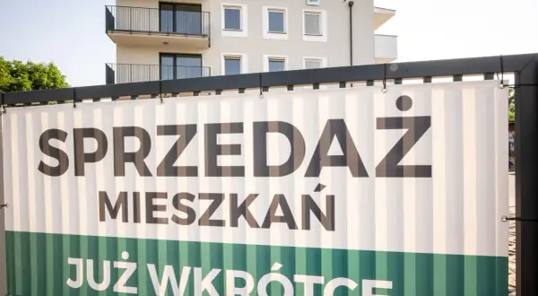 Baner z napisem "SPRZEDAŻ MIESZKAŃ JUŻ WKRÓTCE" na tle nowoczesnego budynku mieszkalnego.